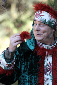 Man wearing a custom santa suit with marijuana leaf print fabric inspects the red fake fur tuft at the end of his green velvet stocking hat